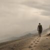 A lone man identified as Sisyphus walks uphill across a barren, rocky slope, leaving footprints in the dusty path. A massive, rough boulder rests beside him on the incline. Hazy mountains stretch across the distance beneath a pale, overcast sky.